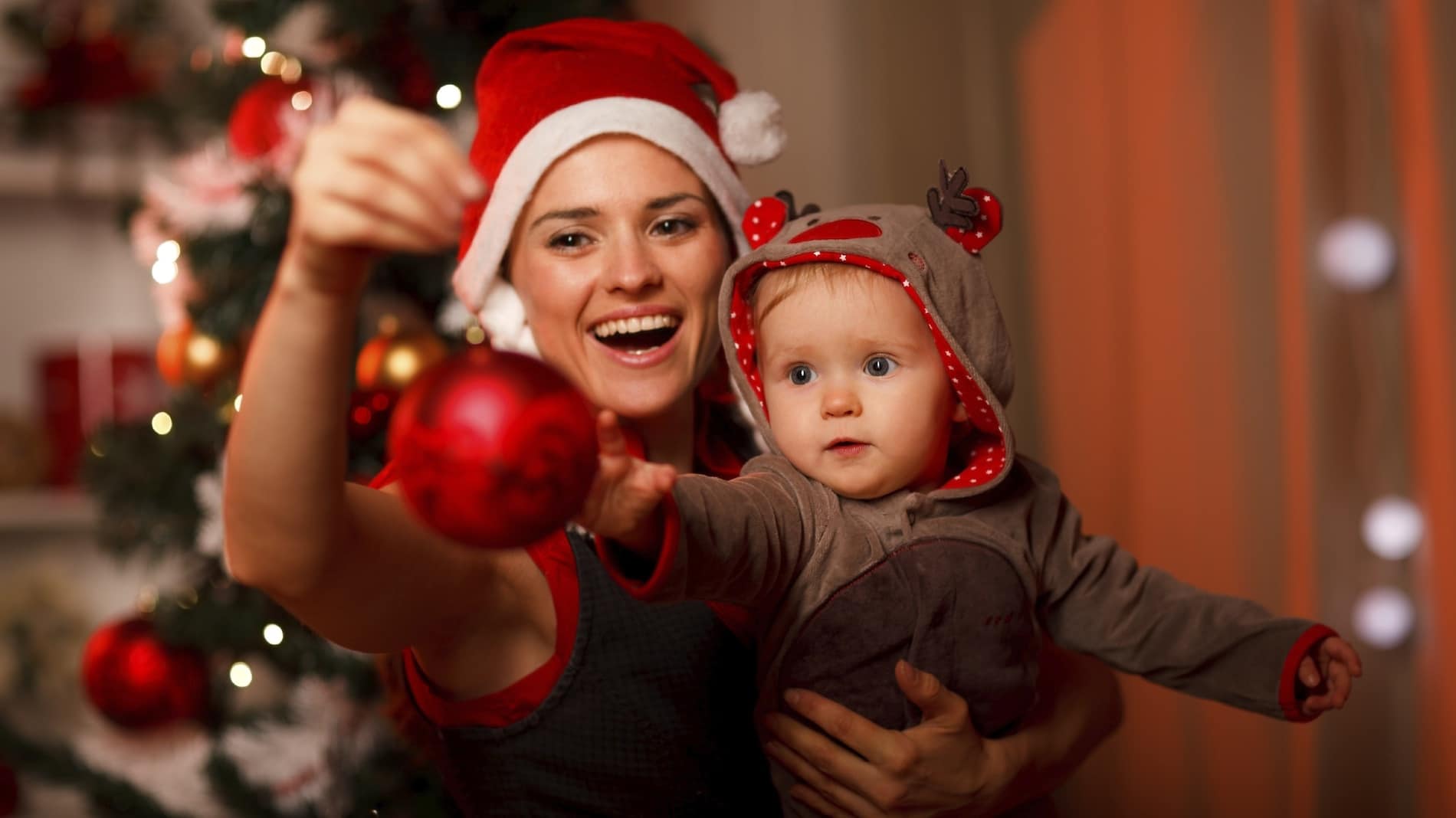 Happy mother showing Christmas ball to baby near Christmas tree