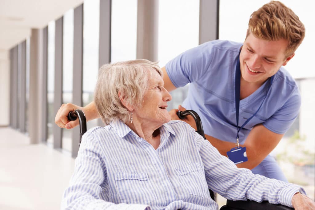 Patient Concierge Pushing Senior Patient In Wheelchair Along Corridor