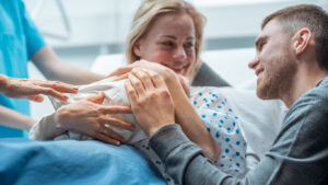 In the Hospital Woman Gives Newborn Baby to a Mother to Hold, Supportive Father Lovingly Hugging Baby and Wife. Happy Family in the Modern Delivery Ward.